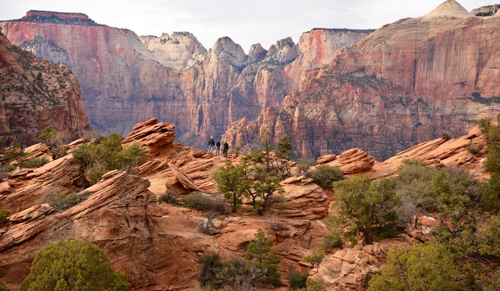 Three hikers stand on layered red sandstone at the Canyon Overlook Trail in Zion National Park, Utah.
