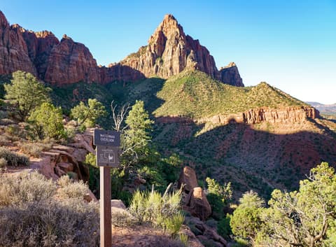 Watchman Overlook trail sign with The Watchman peak and red sandstone canyon walls in Zion National Park.