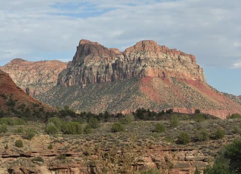 Massive red sandstone butte rises above desert scrub in Zion National Park, Utah, under a partly cloudy sky.