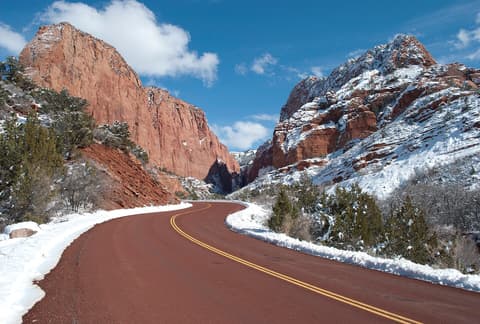 A snow-lined road curves through Kolob Canyon's towering red sandstone cliffs under a bright winter sky.