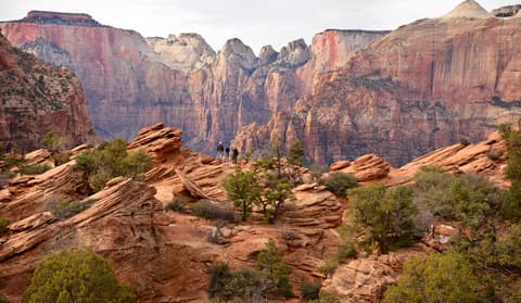 Three hikers stand on layered red sandstone at the Canyon Overlook Trail in Zion National Park, Utah.