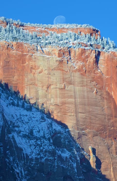A full moon sets behind snow-dusted red sandstone cliffs of Zion National Park in winter.