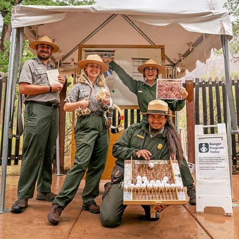 Four smiling Zion National Park rangers pose together at an outdoor Ranger Program Today station in Zion Canyon.