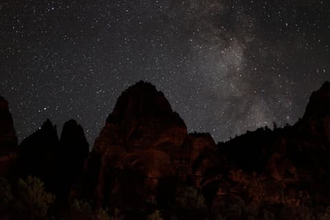 The Milky Way stretches over dramatic red rock canyon silhouettes in Zion National Park, Utah at night.