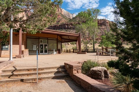 The Zion Human History Museum entrance with towering red sandstone cliffs and blue skies behind it.