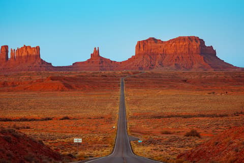 A straight two-lane highway runs through a vast desert landscape toward towering red sandstone buttes and mesas glowing in warm light under a clear blue sky.