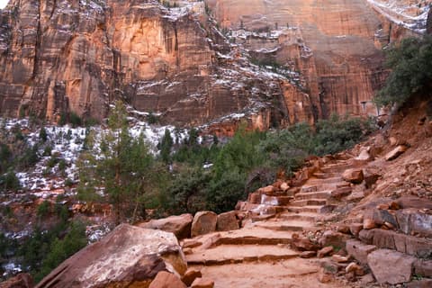 Stone stair steps climb a red sandstone hillside past snow-dusted cliffs and evergreens in Zion National Park.