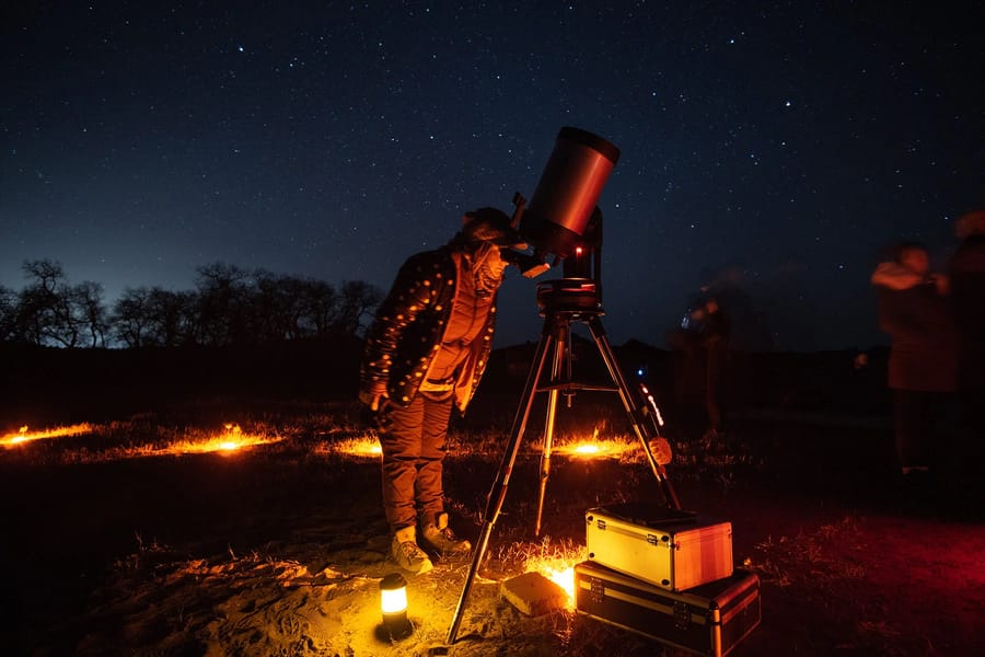 A person in a warm jacket and boots standing next to a telescope under a starry night sky, with a campfire and equipment nearby in a remote, natural setting.