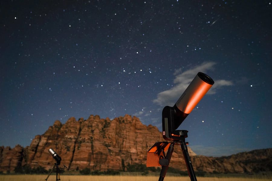 A telescope pointed towards the starry night sky, with towering red rock cliffs of Zion National Park visible in the background.