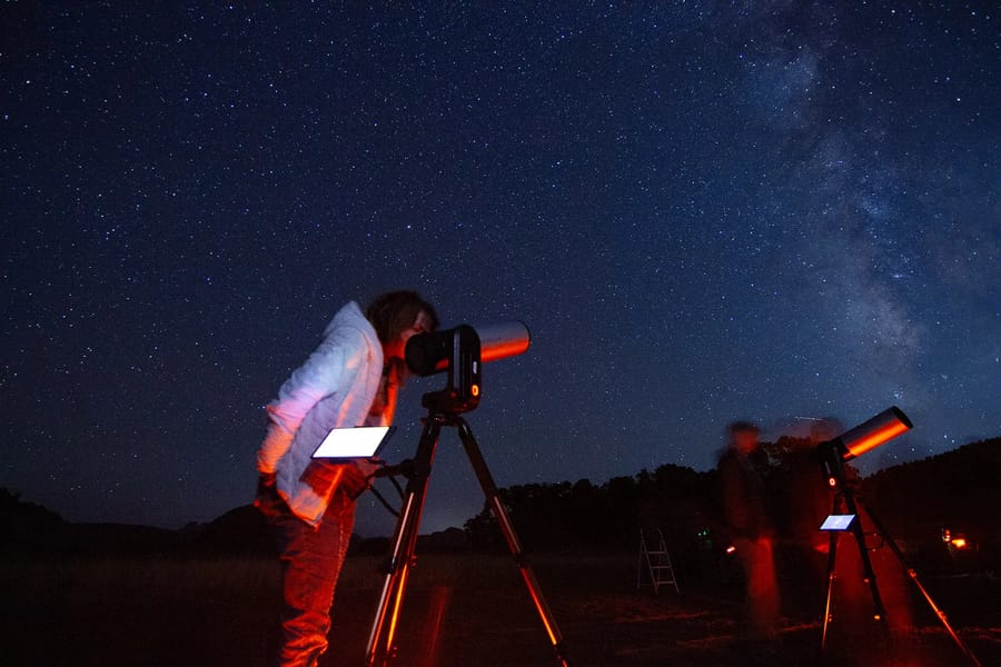 A person in a red jacket stands at a telescope, gazing up at the vast, starry night sky in a remote, mountainous location in southern Utah.