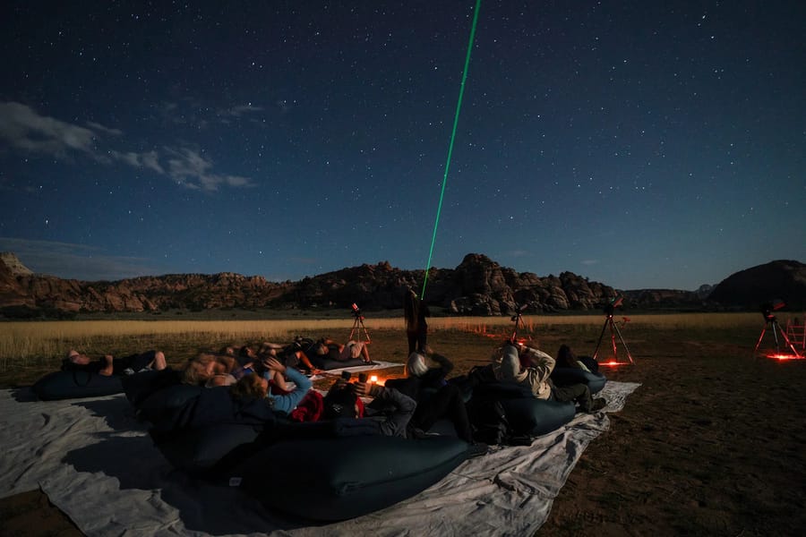 A group of people stargazing under a clear night sky with telescopes and a campfire in Zion National Park.